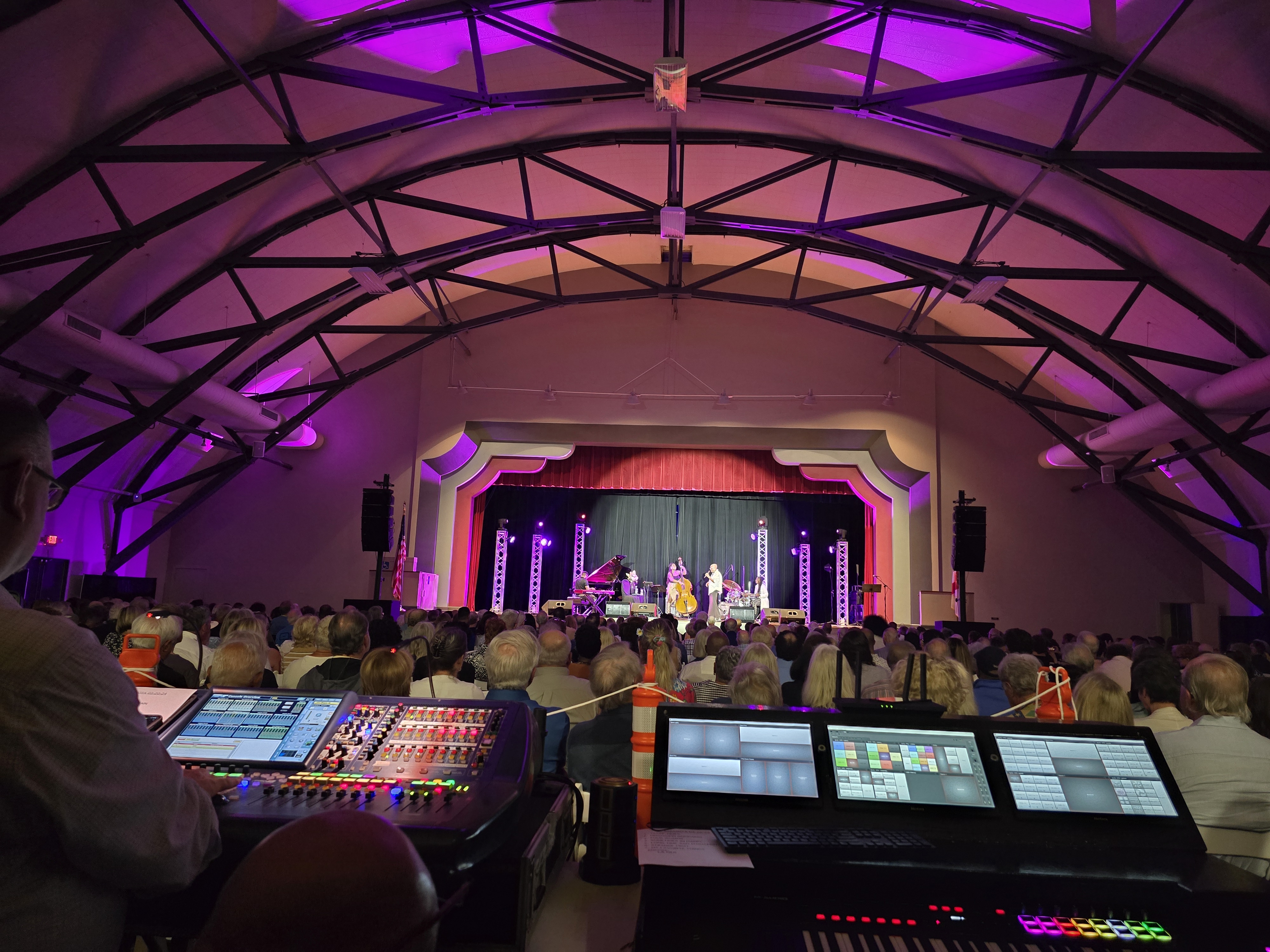 Sound and lighting consoles overlooking a live band and seated audience beneath a purple-lit arched ceiling